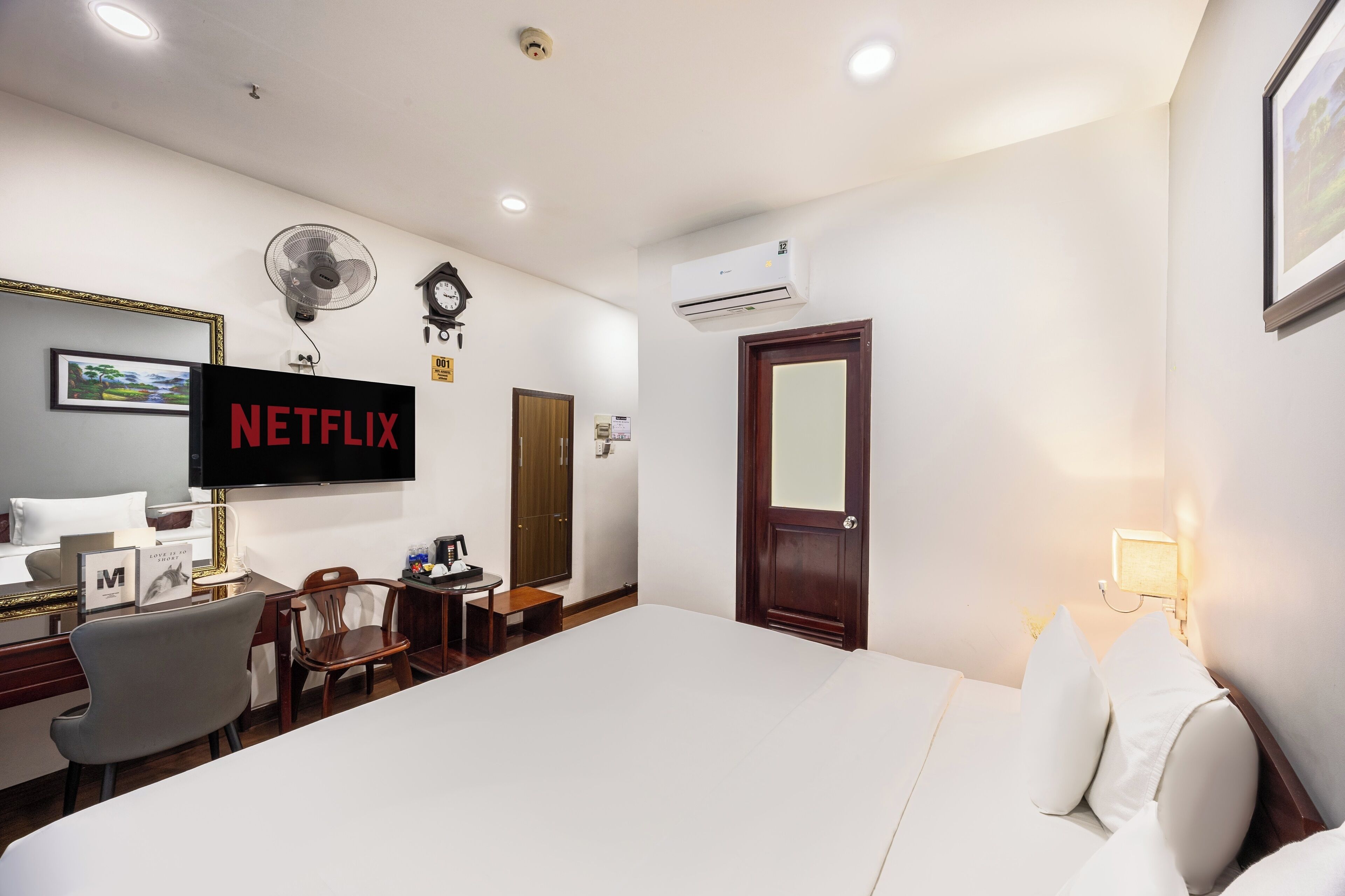 A hotel room with a bed, a desk with a laptop, a chair, a TV mounted on the wall, and an air conditioning unit.