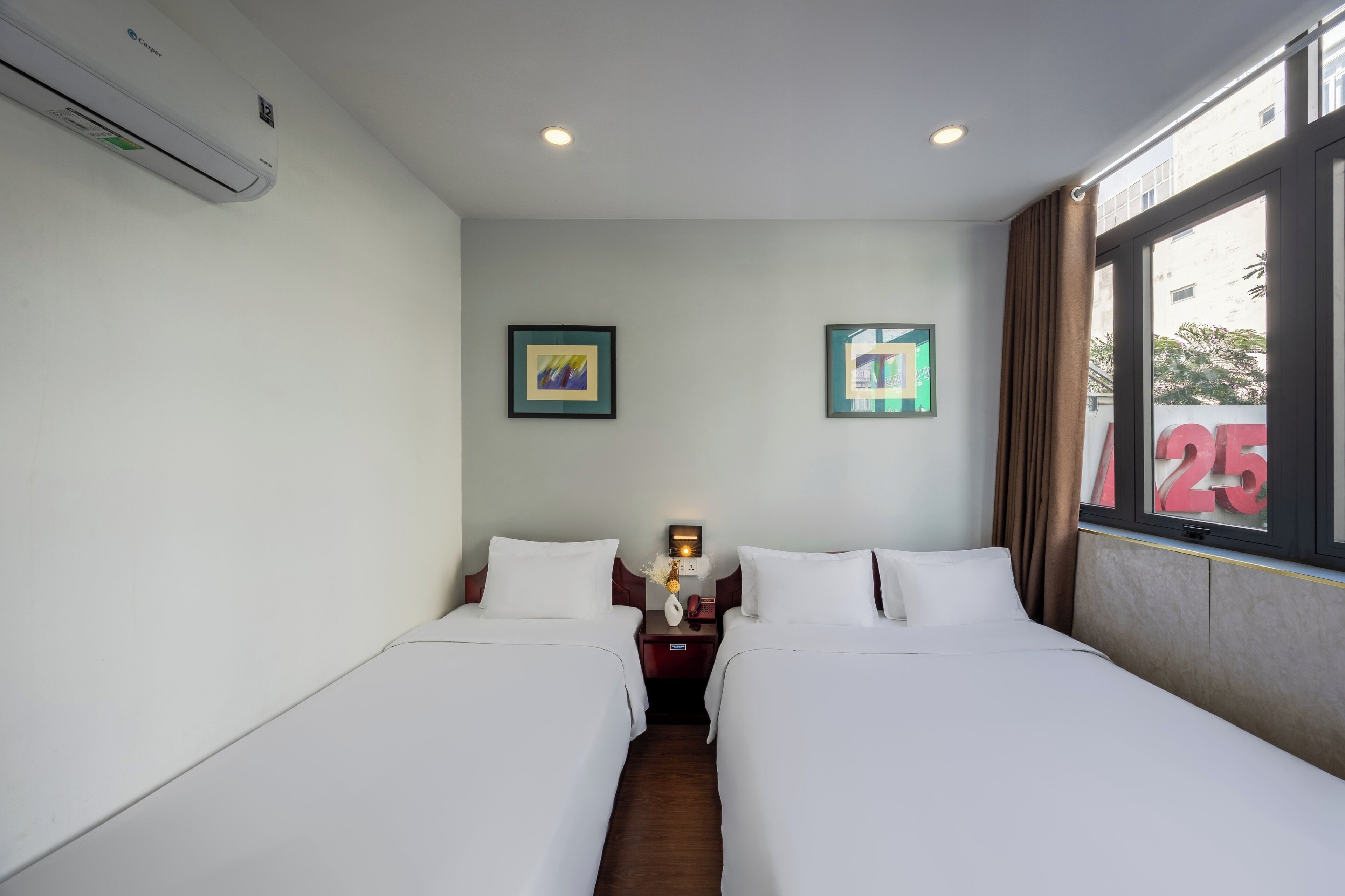 A hotel room with two beds, a nightstand with a lamp, framed pictures on the wall, and a window with curtains.