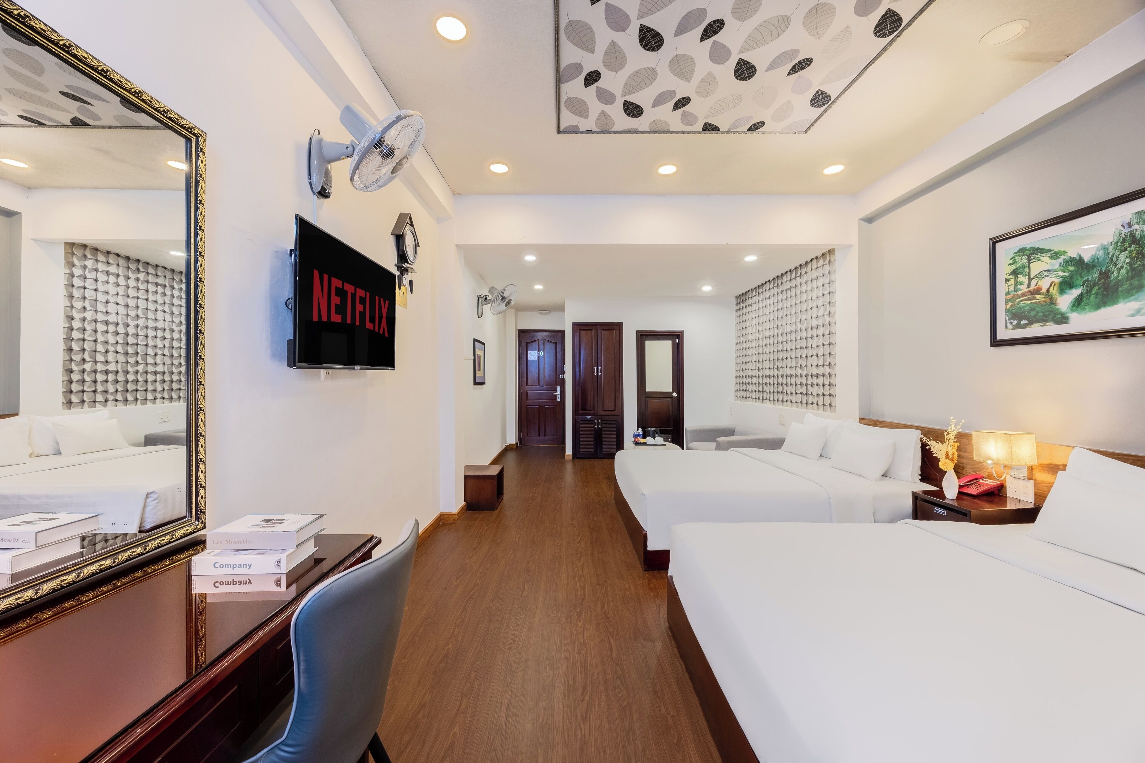 Habitación de hotel con un espejo grande, un televisor de pantalla plana, dos camas, un escritorio con una silla y un ventilador de techo.
