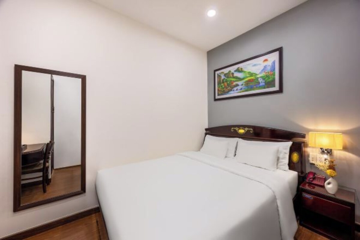 A hotel room with a bed, a nightstand with a lamp, a painting on the wall, and a mirror on the wall.