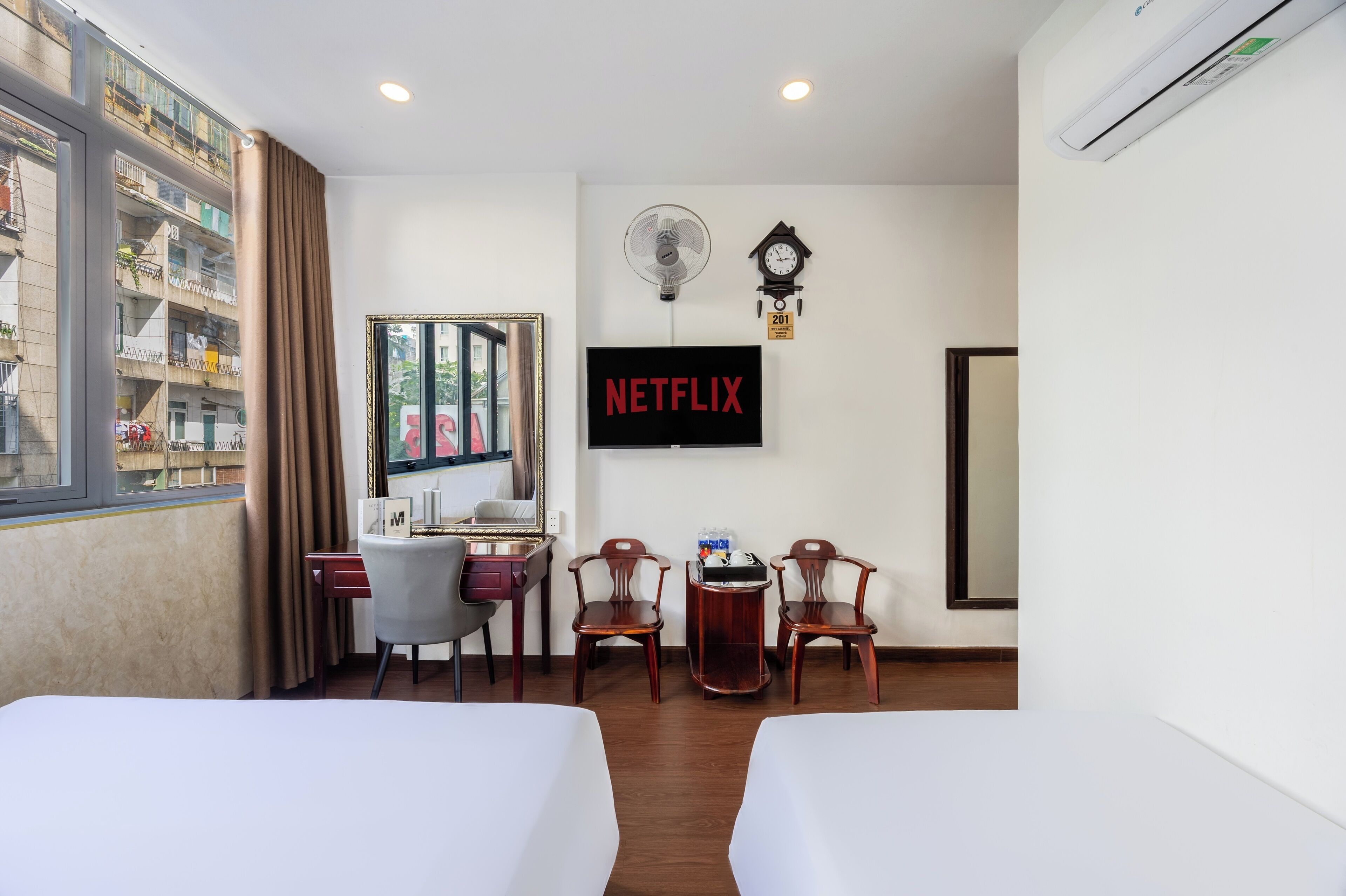 A hotel room with two beds, a desk with a chair, a small table with a chair, a wall-mounted TV, a clock, and an air conditioner.