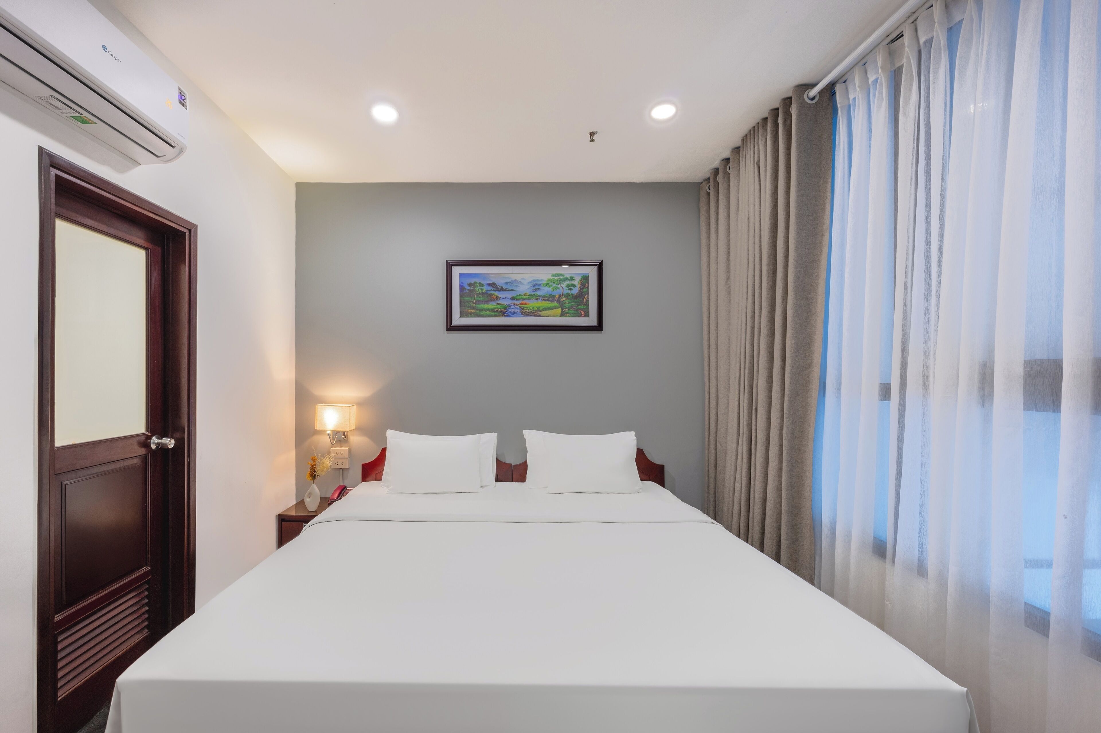 A hotel room with a bed, a nightstand with a lamp, a framed picture on the wall, and a window with curtains.