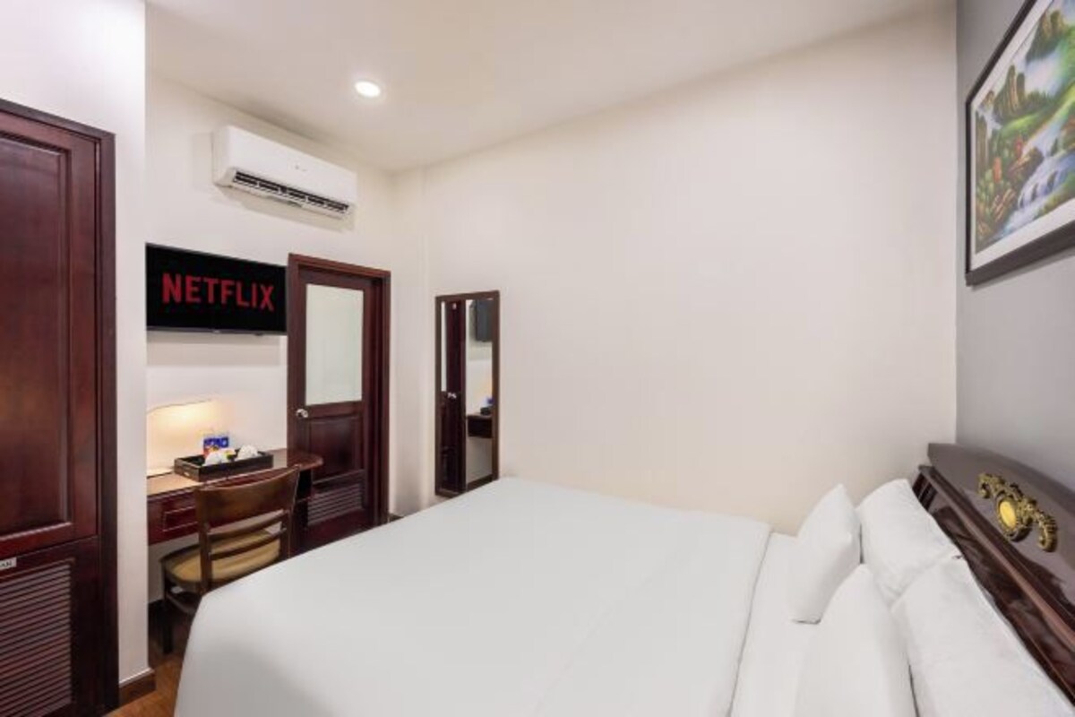 A hotel room with a bed, a desk with a chair, an air conditioning unit, a door, and a wall-mounted sign that reads "NETFLIX".