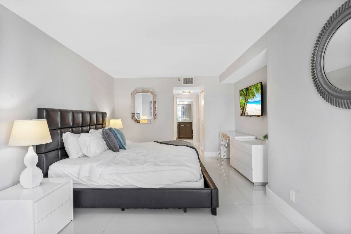 A modern bedroom with a large bed, bedside tables, a mirror, a TV, and a bathroom with a sink and mirror.