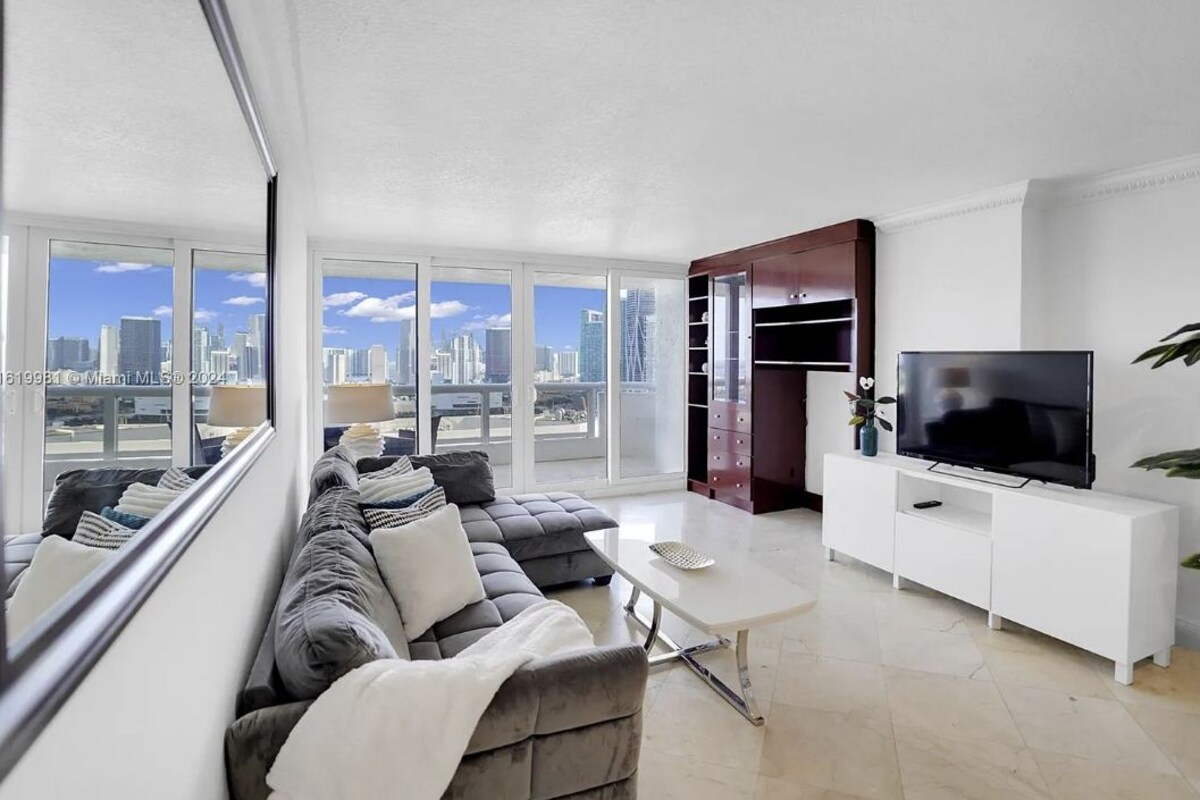 Un salon moderne avec un grand canapé, une table basse, un téléviseur posé sur un meuble blanc et une vue sur la ville à travers de grandes fenêtres.