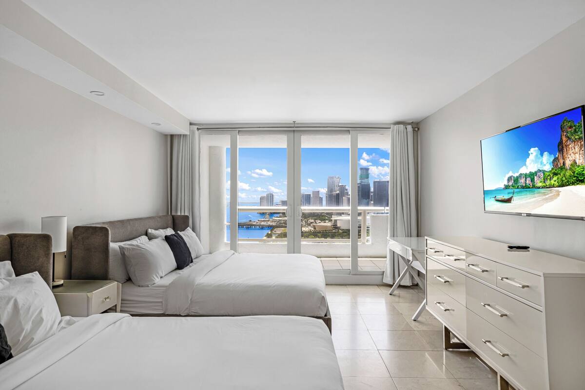 A hotel room with two beds, a desk, a television, and a large window with a city and water view.