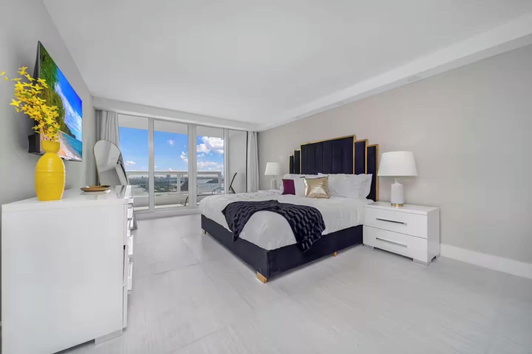 A modern bedroom with a large bed, bedside tables, a desk, and a city view.