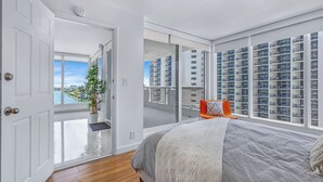 Een moderne slaapkamer met een groot bed, uitzicht op de stad door middel van ramen van vloer tot plafond, en een balkon met een hekwerk.