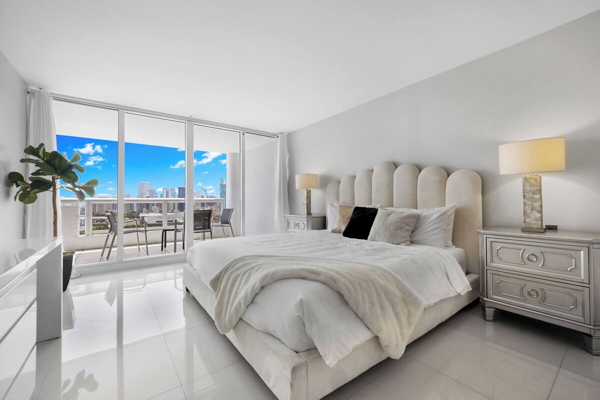A modern hotel room with a large bed, bedside lamps, a nightstand, and a view of the city.