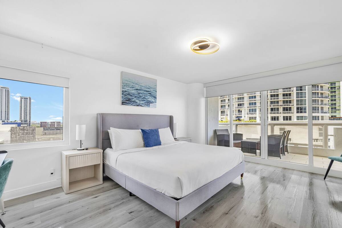 A modern hotel room with a large bed, a nightstand, a chair, a window with a city view, and a balcony with outdoor seating.