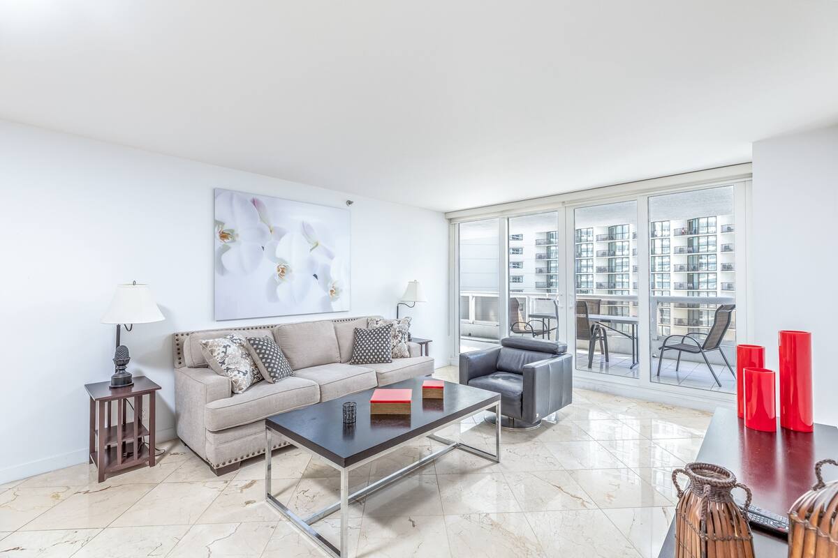 A modern living room with a sofa, coffee table, and chairs, a large window with a view of high-rise buildings, and a painting on the wall.