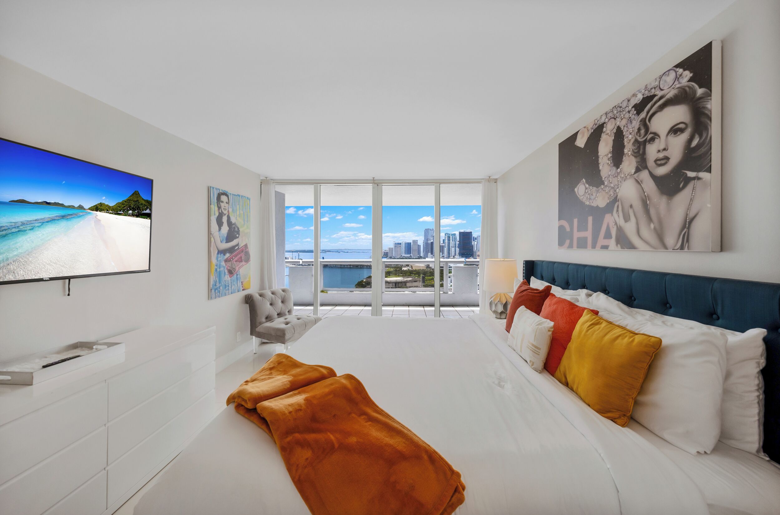 A modern hotel room with a large TV, a bed with colorful pillows, a sofa, and a city view.