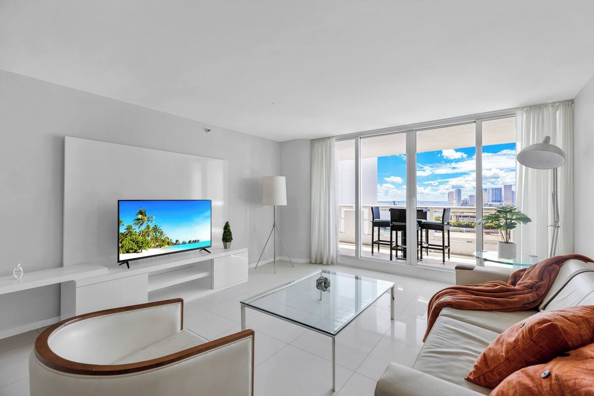 A modern living room with a glass coffee table, a sofa, a TV on a white stand, and a large window with a city view.