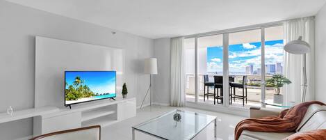 A modern living room with a glass coffee table, a sofa, a TV on a white stand, and a large window with a city view.