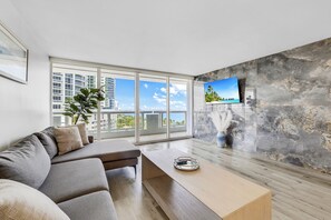 A modern living room with a large window, a grey sofa, a wooden coffee table, and a mounted TV. - Comfort, Space and Perfect Sunset (Miami)