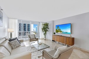 A modern living room with a sofa, armchairs, a coffee table, a TV, and a large window with a city view.