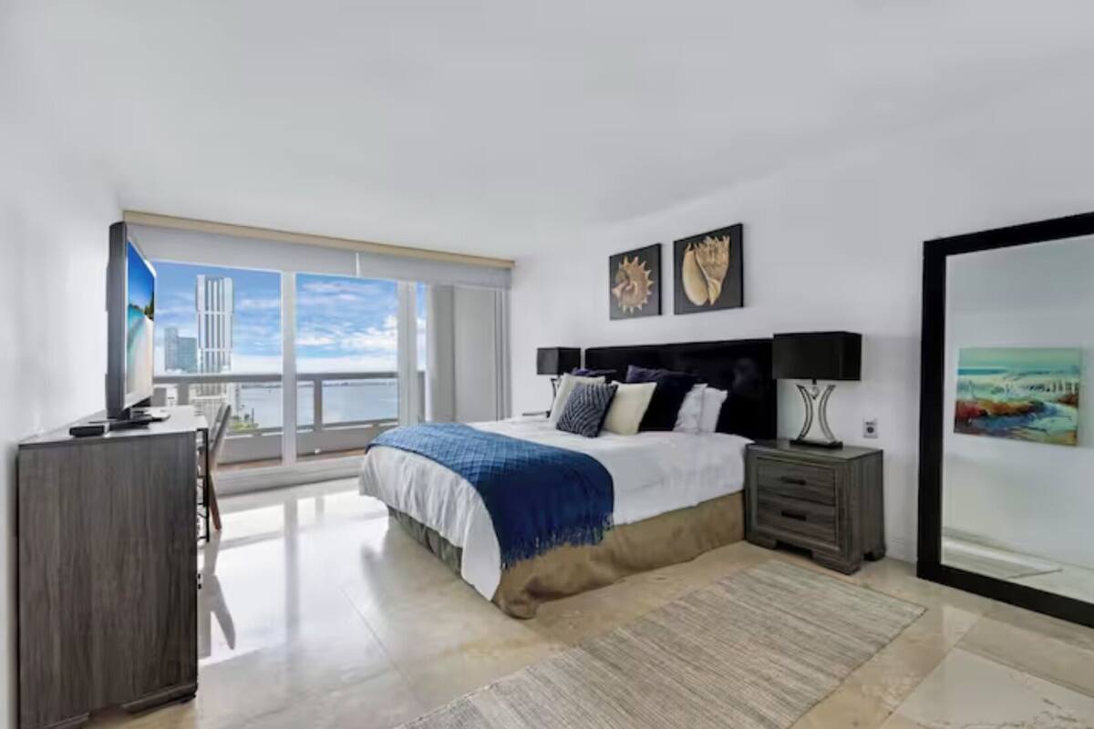 A modern bedroom with a large bed, a TV, a desk, and a view of the city.