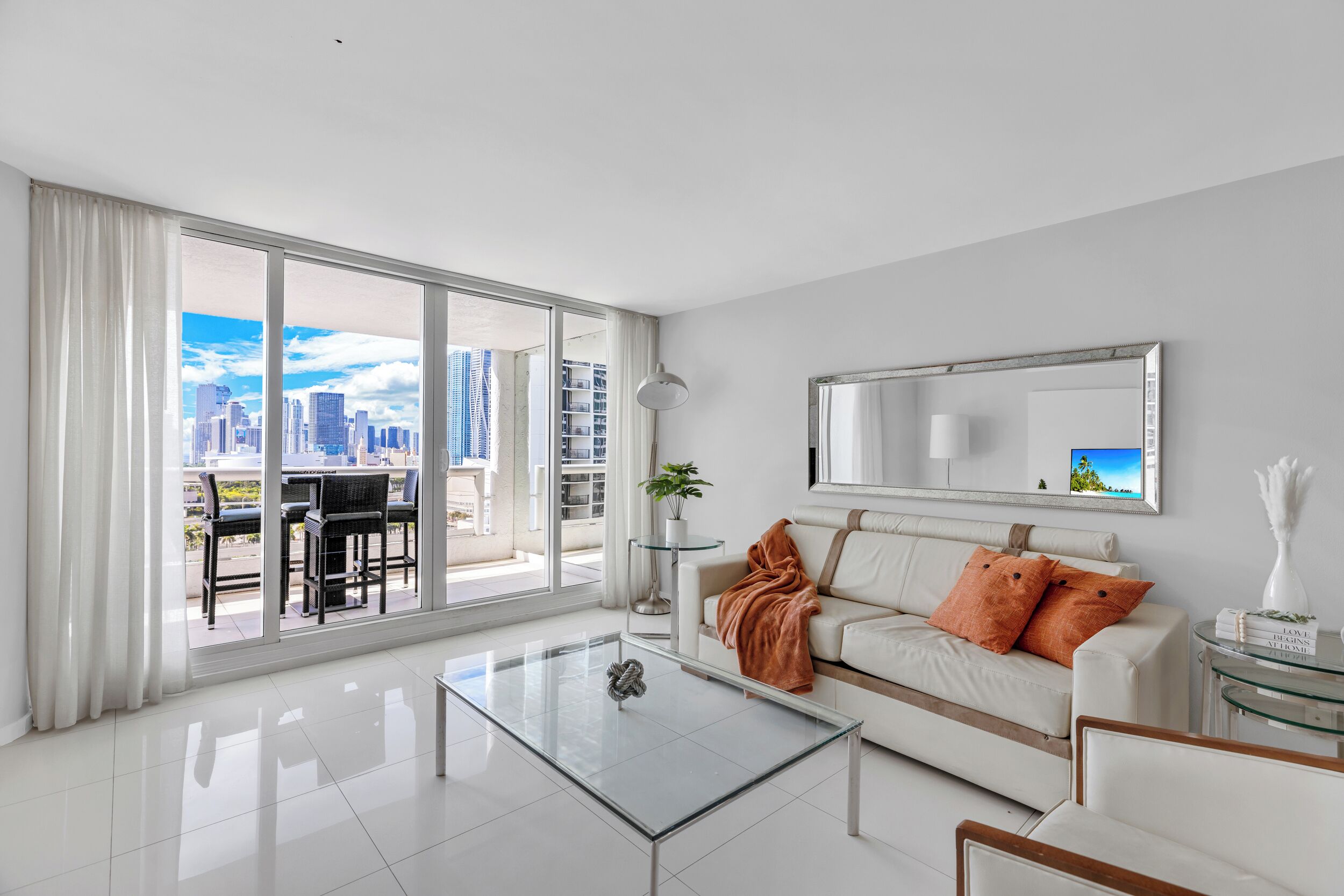 A modern living room with a glass coffee table, white sofa, and large windows offering a city view.