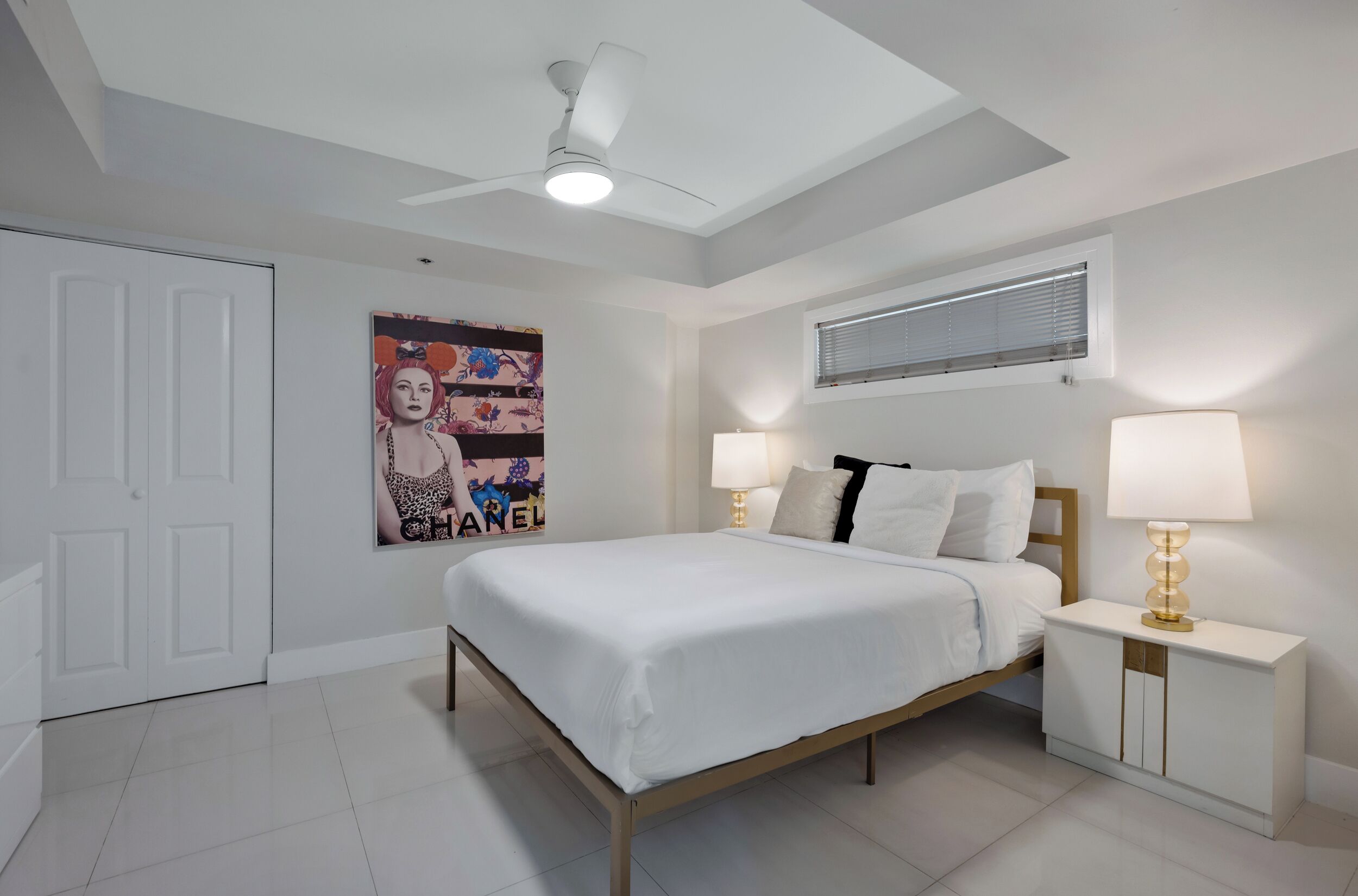 A bedroom with a bed, two bedside lamps, a ceiling fan, a poster on the wall, and a window with blinds.
