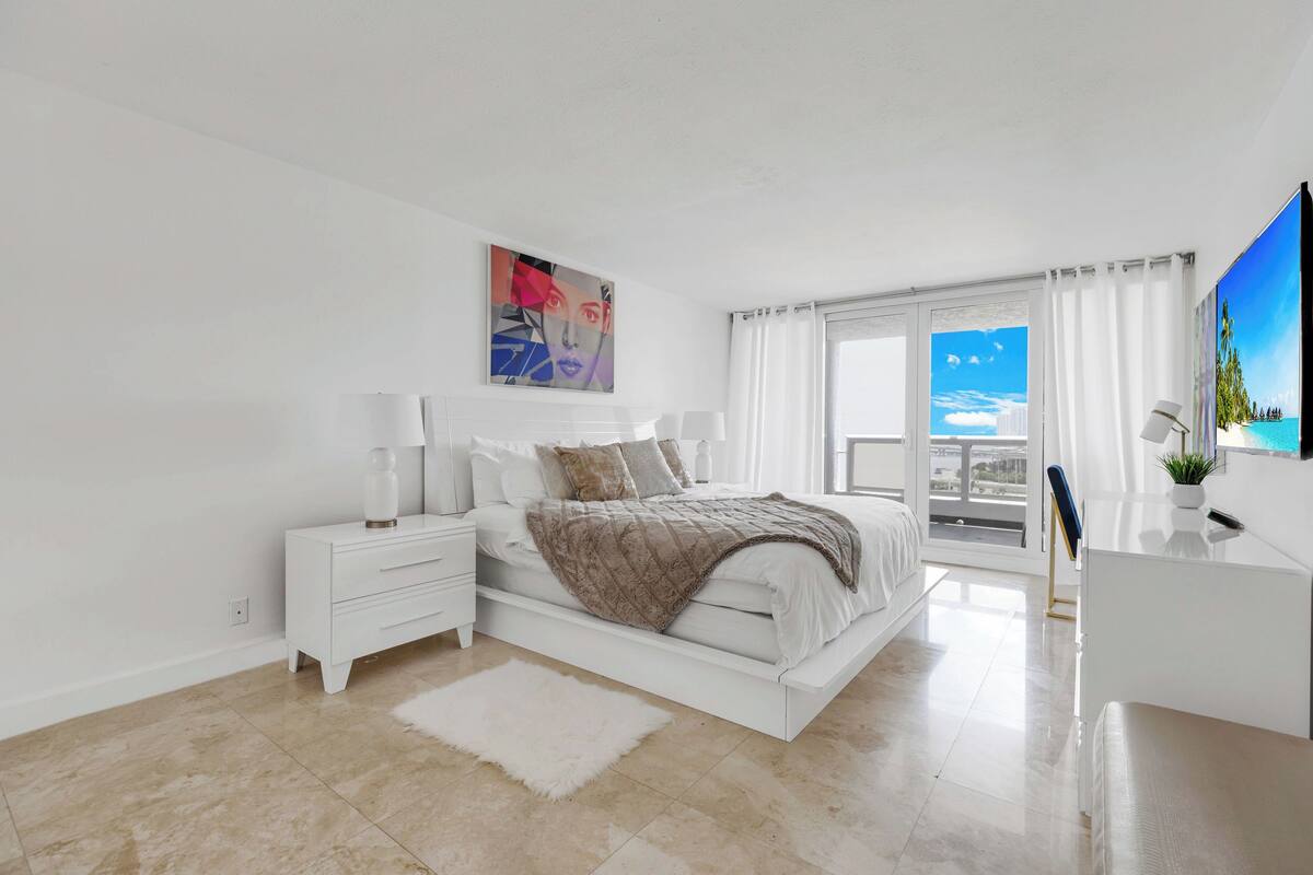 A modern bedroom with a large bed, white nightstands, a desk, and a view of the sky through a window.