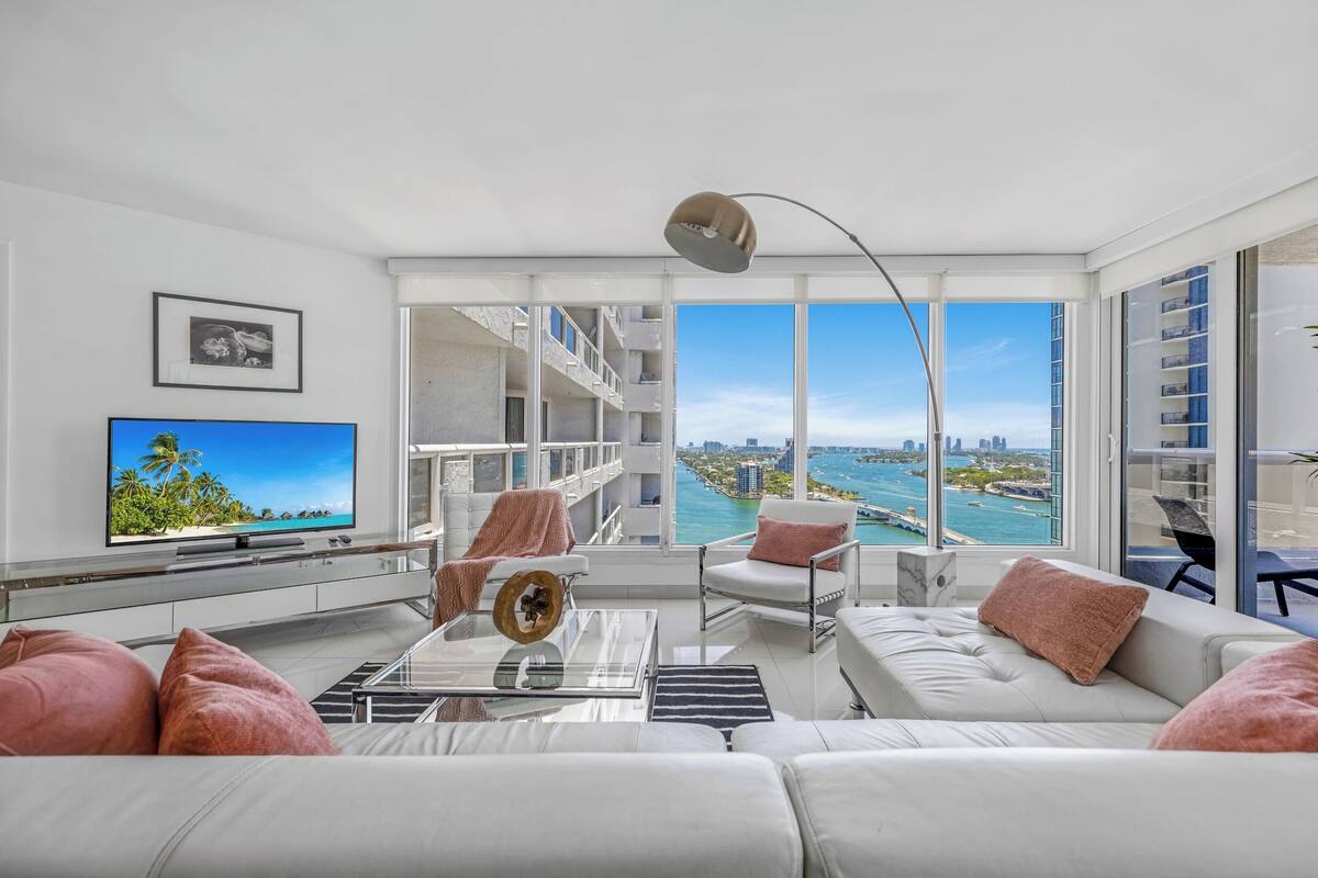 A modern living room with a large TV, comfortable seating, a coffee table, and a view of a city and water.