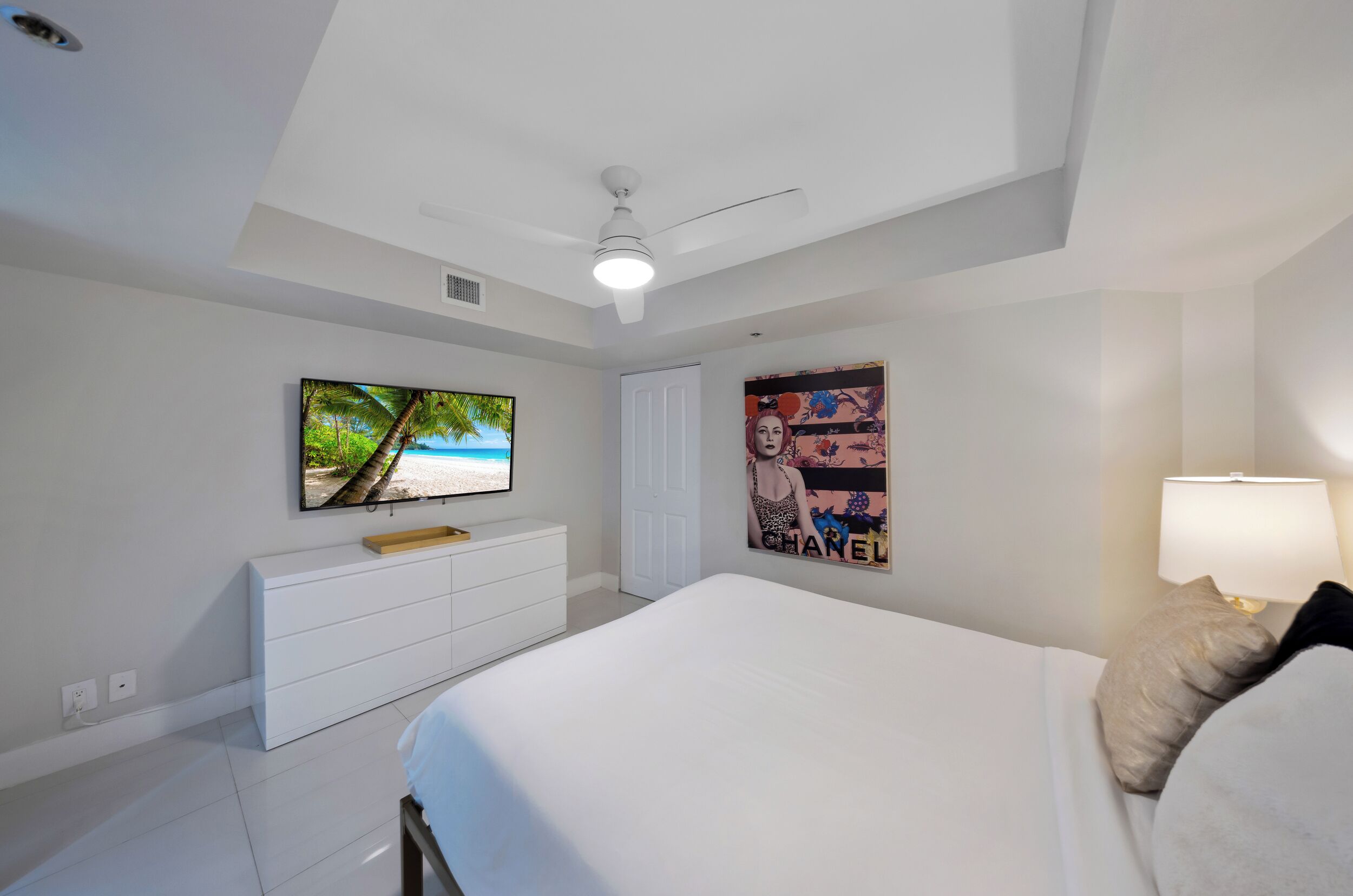 Un dormitorio con cama, televisor montado en la pared, ventilador de techo, cómoda y un cartel.