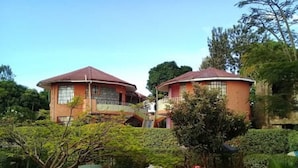 A two-story building with a red roof, surrounded by lush greenery and a garden with outdoor furniture. - Jambo Village Hotel (Murang'a)