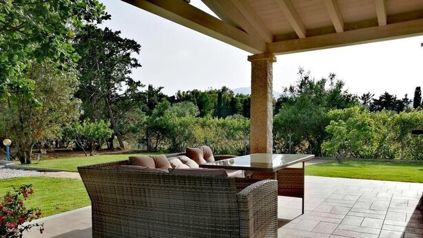 A covered outdoor seating area with a wicker sofa and a table, surrounded by a landscaped garden and trees.