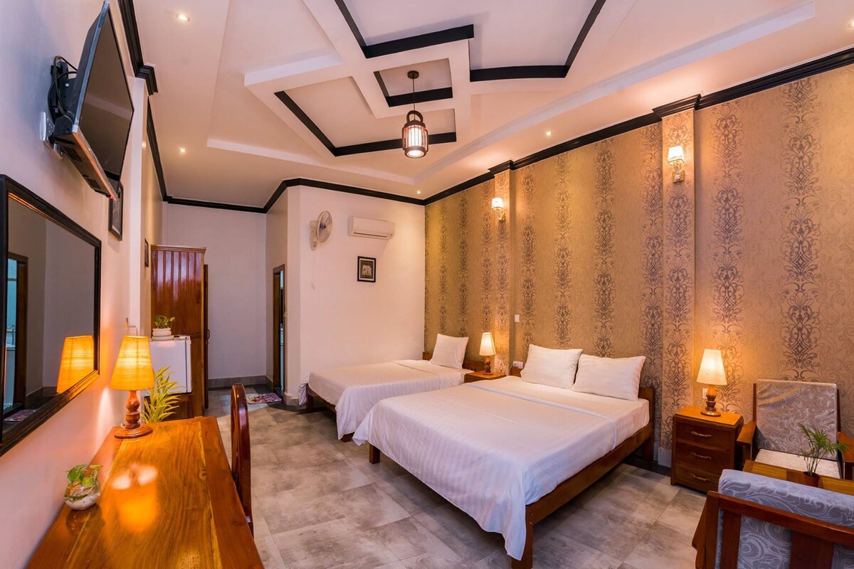 A hotel room with two beds, a wooden dining table, and a TV mounted on the wall.