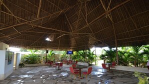 Un patio techado al aire libre con suelo de piedra, sillas y mesas rojas, rodeado de follaje.