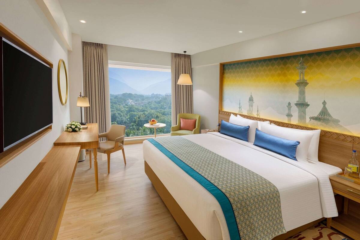 Ein Hotelzimmer mit einem großen Bett, einem Schreibtisch, einem Sessel, einem kleinen Tisch mit Blumenvase und Bergblick durch das Fenster.