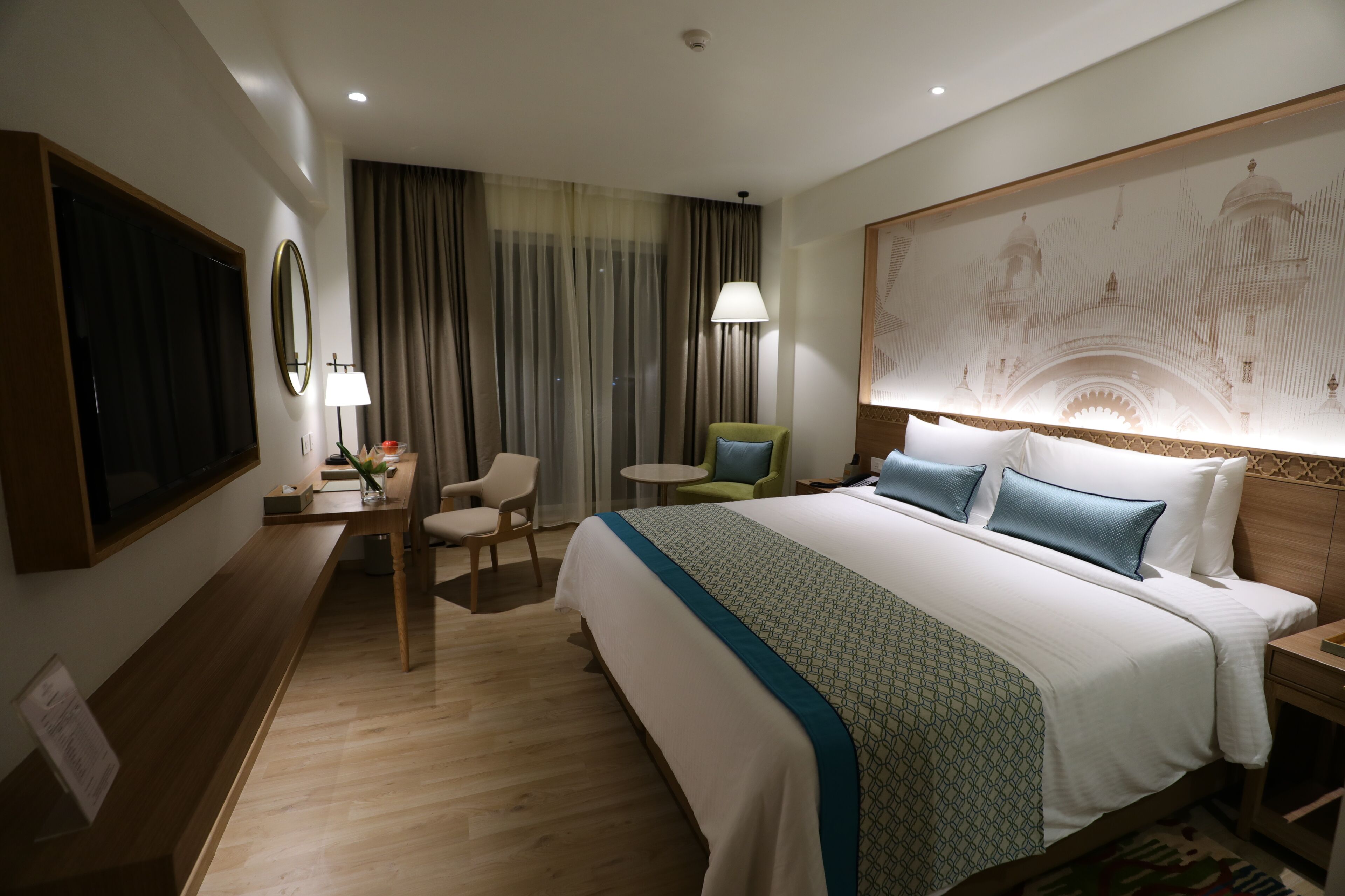 Quarto de hotel com uma cama grande, uma escrivaninha com cadeira, televisão e um quadro grande na parede.