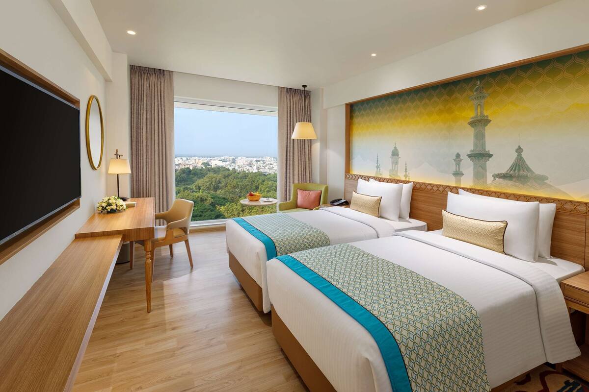 Ein Hotelzimmer mit zwei Betten, einem Schreibtisch, einem Sessel, einem Fernseher und einem großen Fenster mit Blick auf die Stadt.