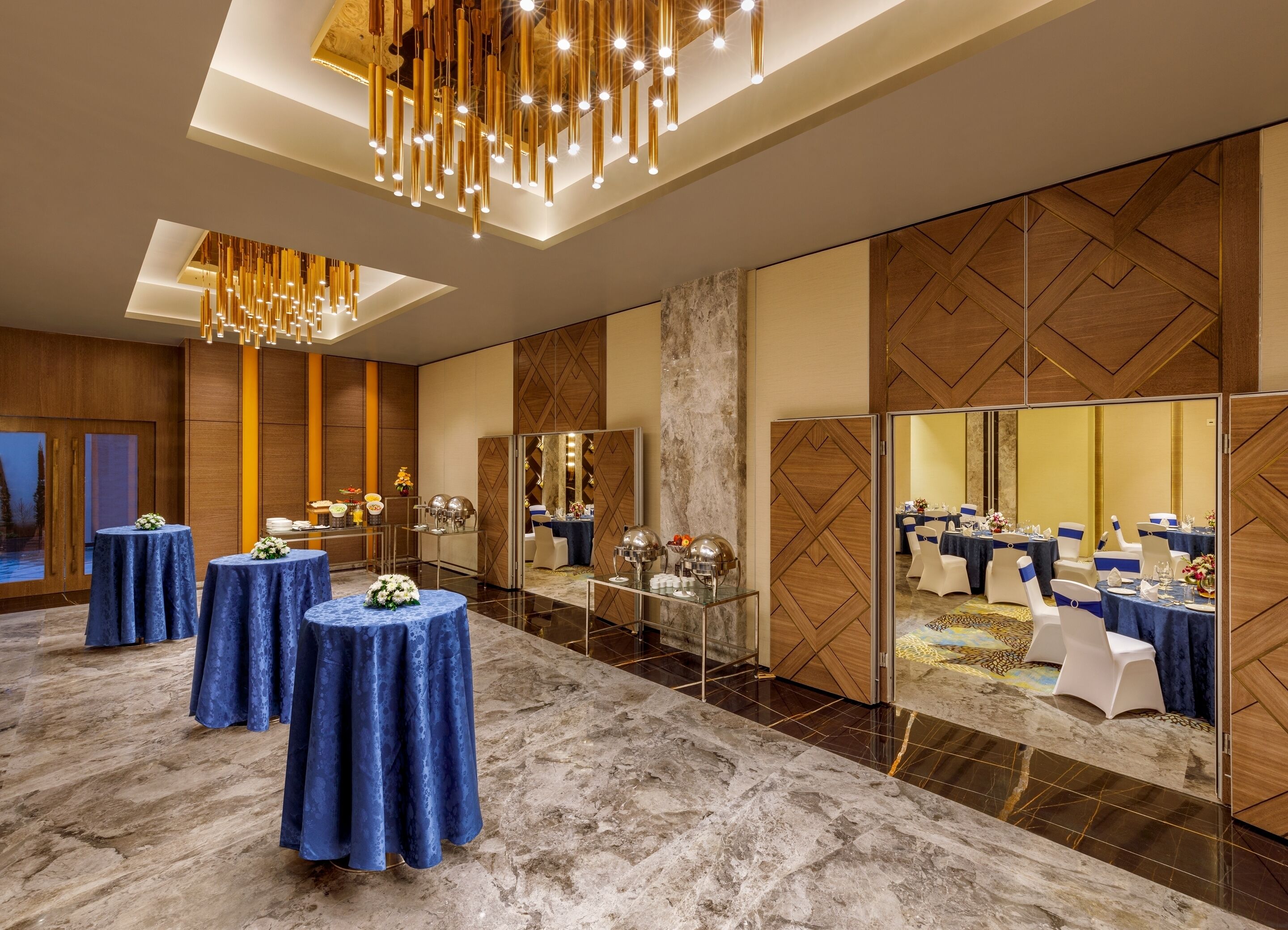 A modern hotel lobby with marble flooring, round tables with blue tablecloths, and a large chandelier.