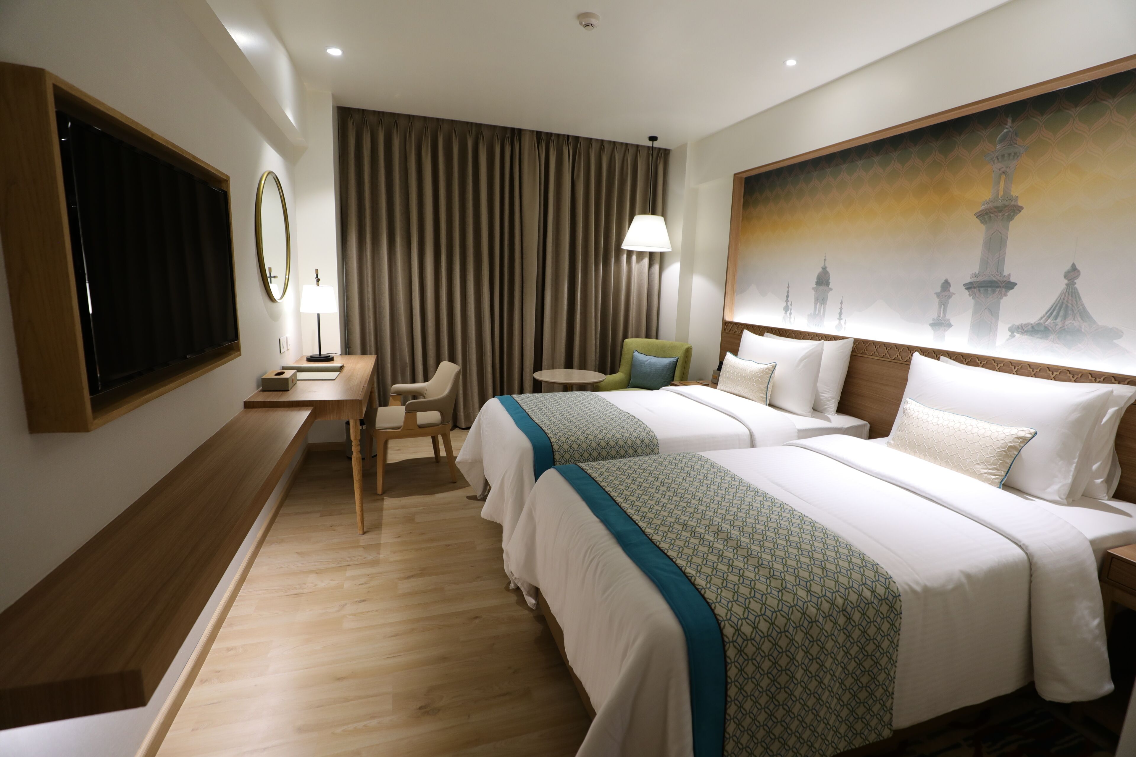 A hotel room with two beds, a desk, a chair, a TV, and a painting of a mosque.