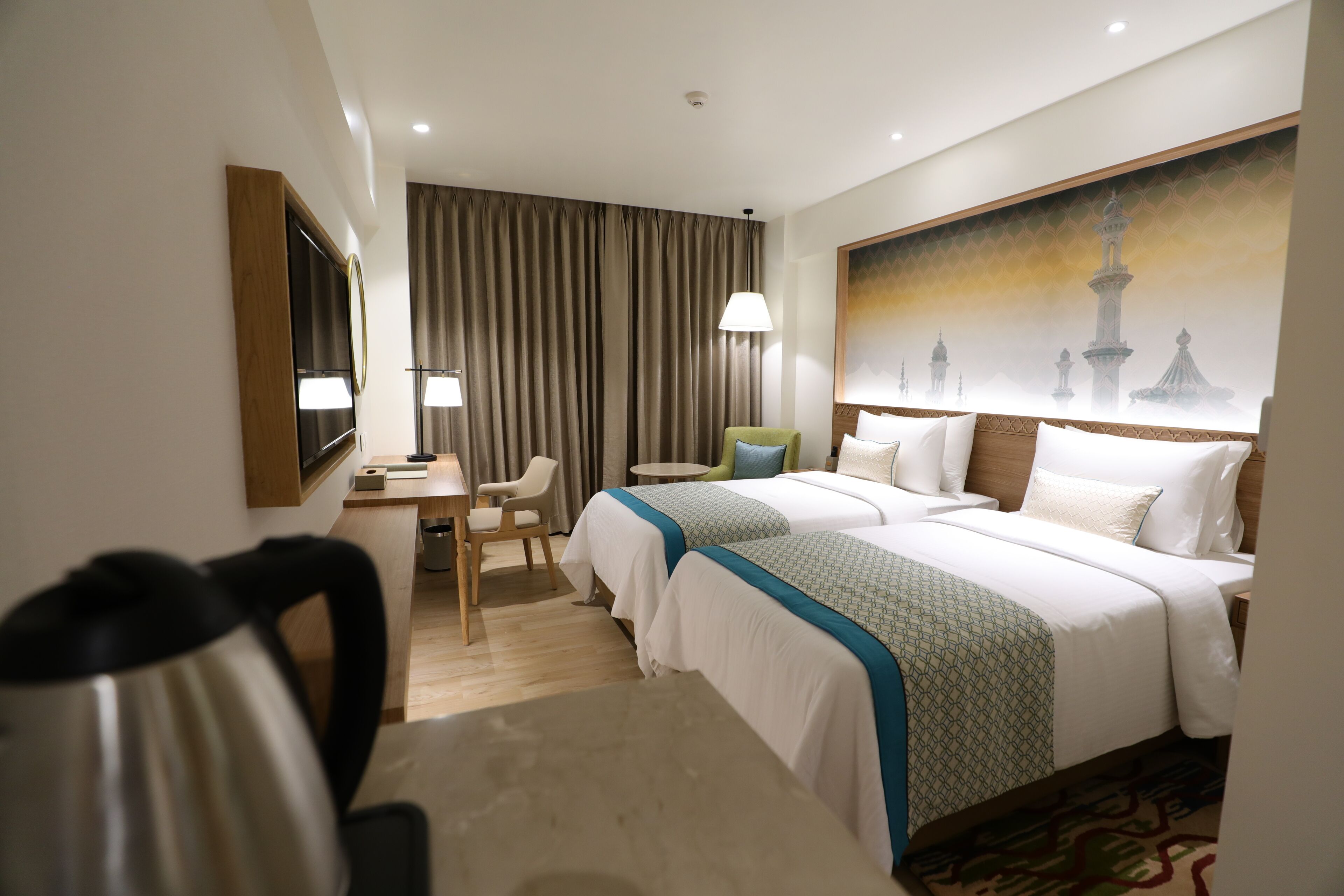 Habitación de hotel con dos camas, un escritorio, una silla, una lámpara y un cuadro de una mezquita.