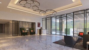 A spacious lobby with a marble floor, a reception desk, two green armchairs, a modern chandelier, and large windows offering a view of greenery.