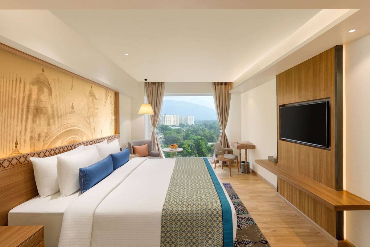 Ein Hotelzimmer mit einem großen Bett, einem an der Wand montierten Fernseher, einem Schreibtisch mit Stuhl und Blick auf die Stadtlandschaft durch das Fenster.