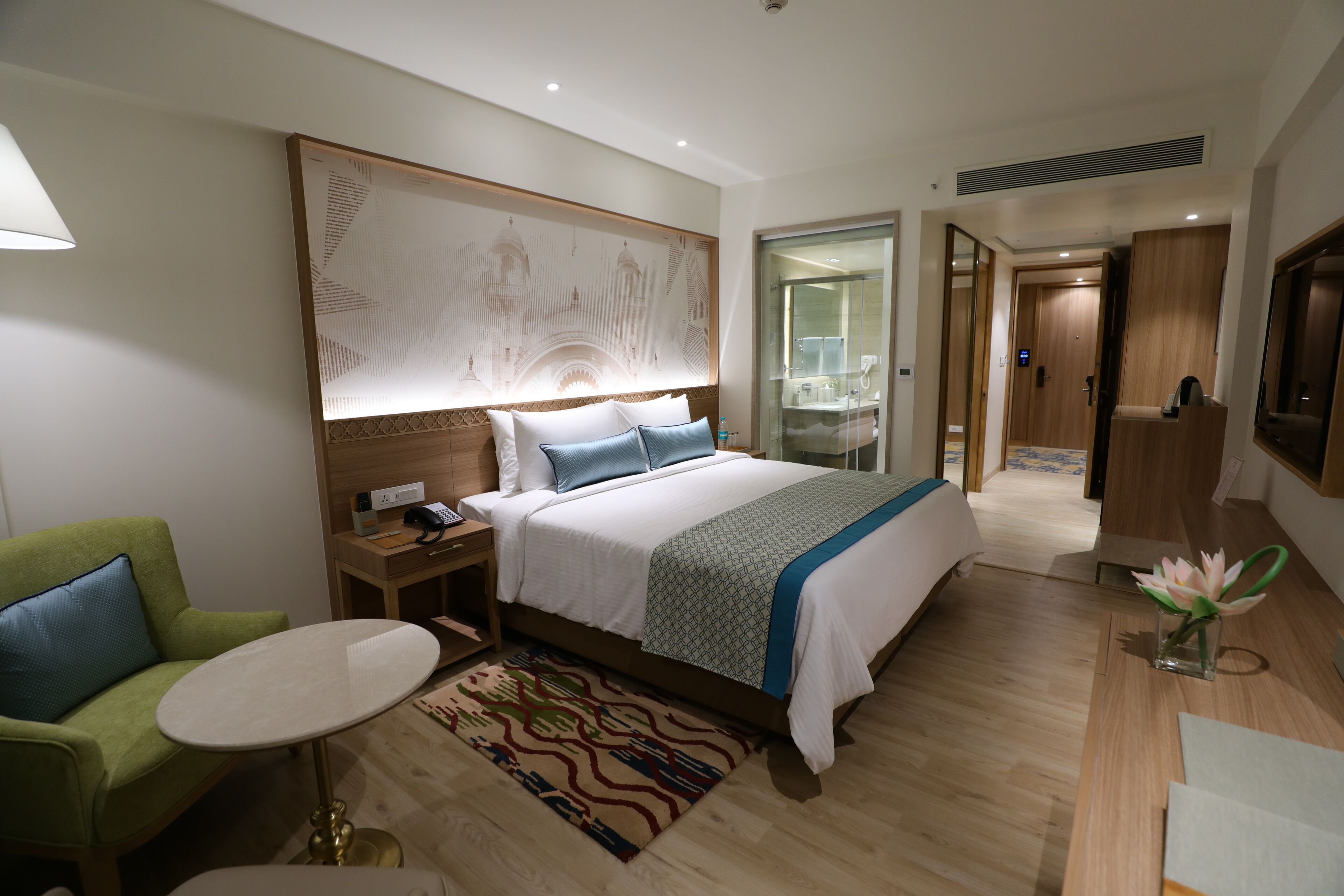 A hotel room with a bed, a green armchair, a small table, a decorative wall piece, and a bathroom visible through an open door.