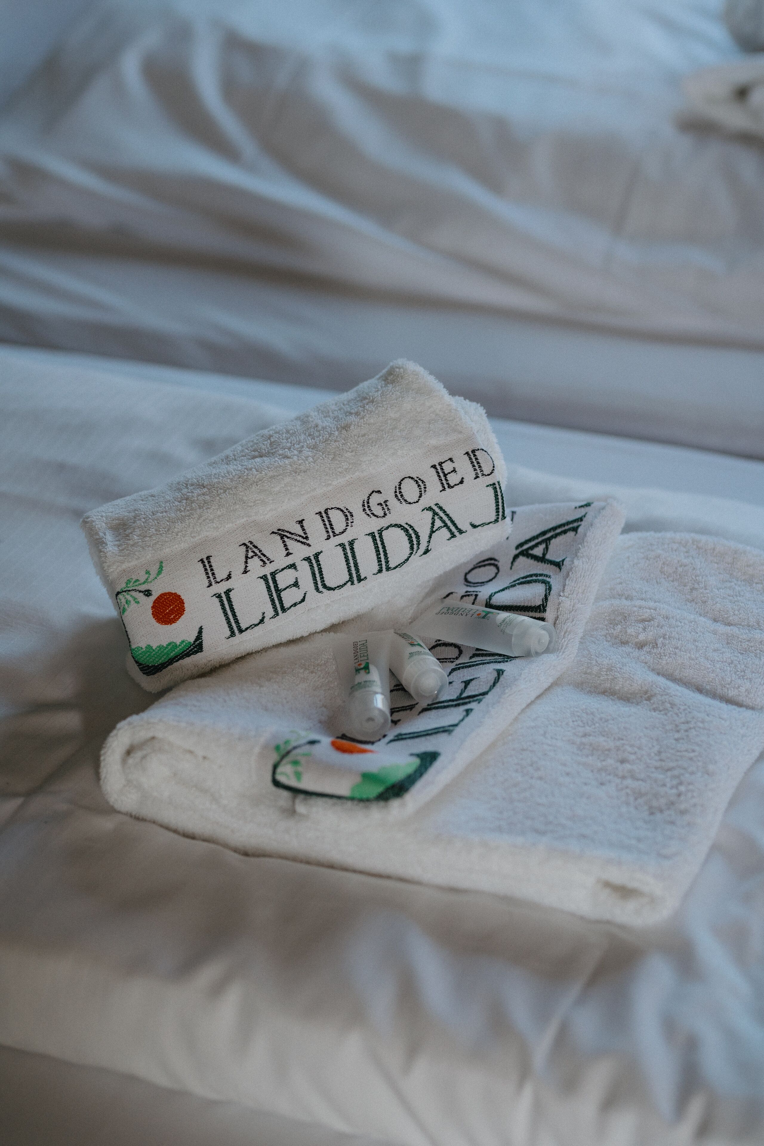 Ein ordentlich gefaltetes Handtuch mit dem Schriftzug "LANDGOED LEUDAJ" und ein Paar Hausschuhe auf einem Bett.