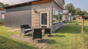 A wooden cabin with a large window, a patio with two chairs, and a small shed in the background. - Landgoed Leudal (Haelen)