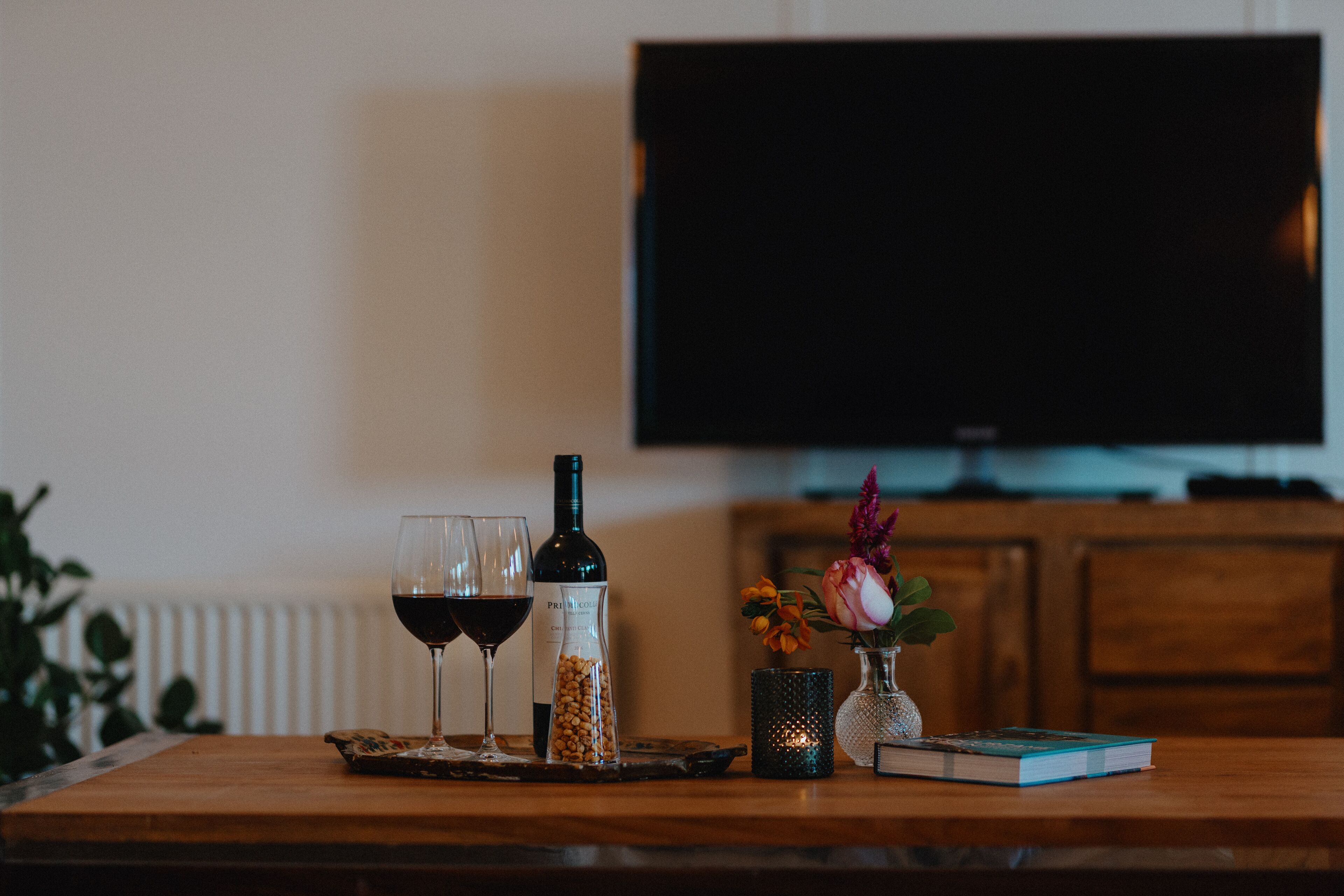 A wooden table with a bottle of wine, two wine glasses, a book, a small vase with flowers, and a candle.