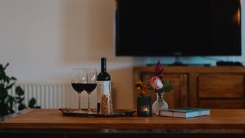 A wooden table with a bottle of wine, two wine glasses, a book, a small vase with flowers, and a candle.