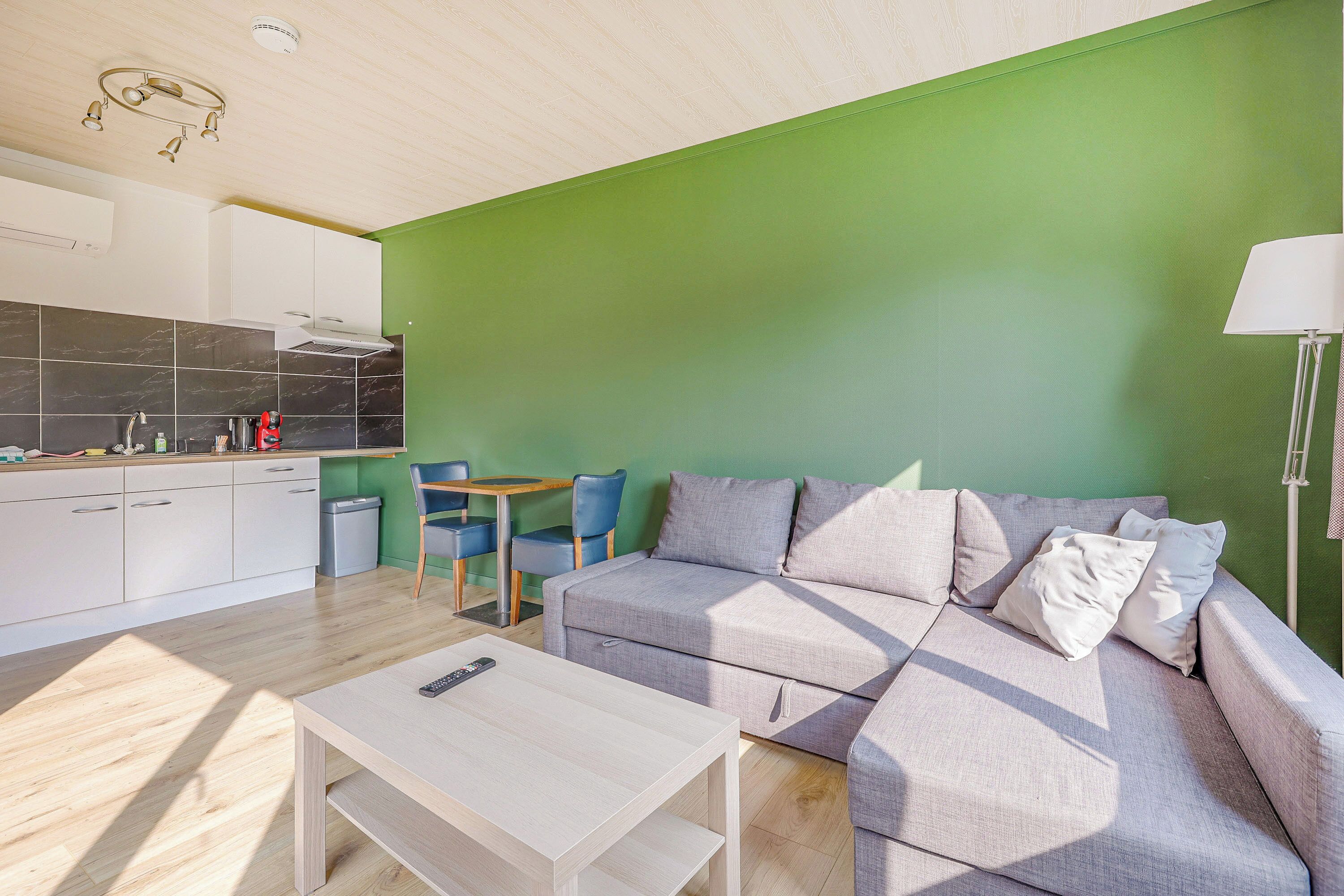 A modern living room with a green accent wall, a grey sofa, a wooden coffee table, and a kitchen area with white cabinets and a black backsplash.