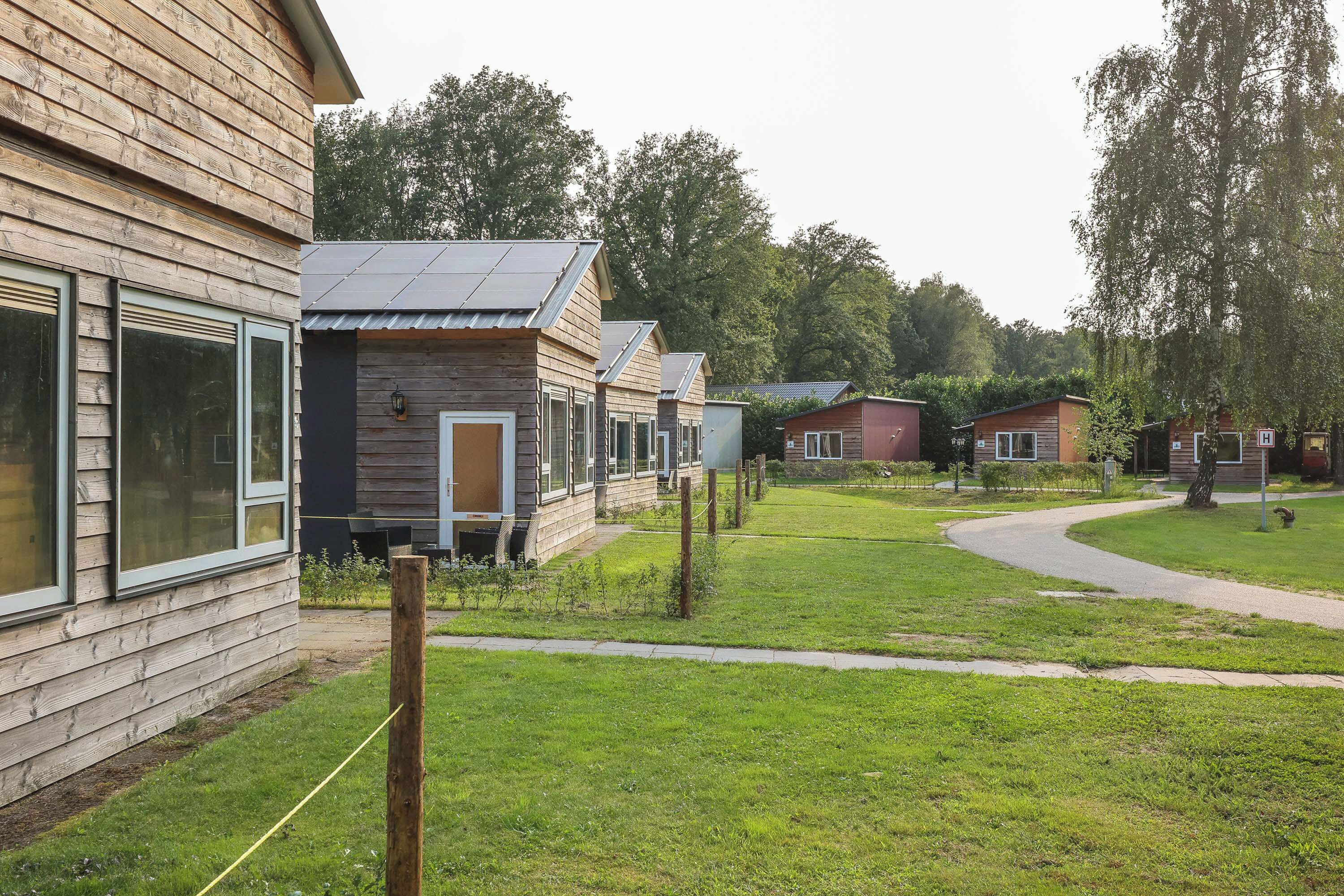A row of wooden cabins with large windows and solar panels, surrounded by a grassy area and trees.