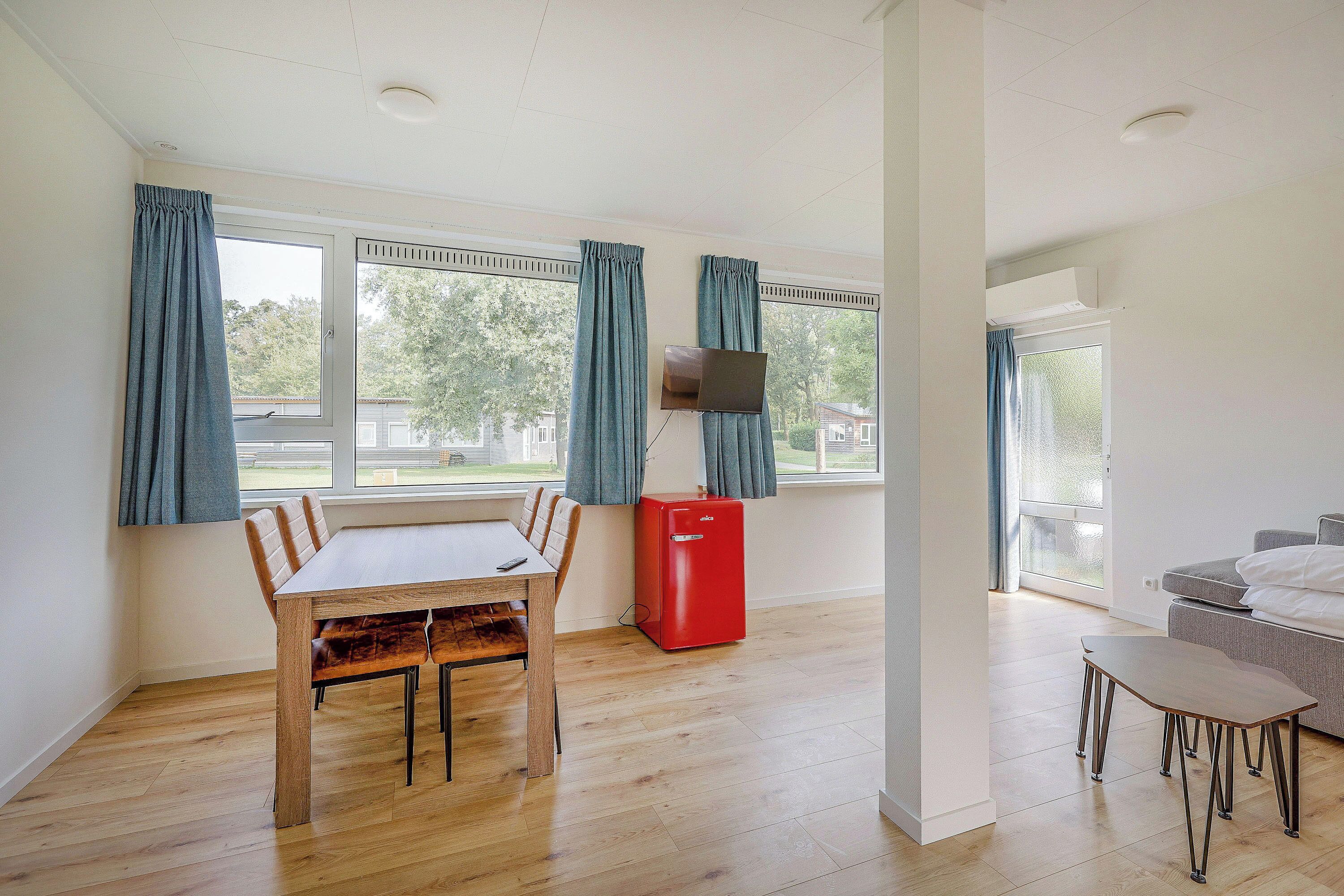Een kamer met een houten eettafel, stoelen, een rode koelkast, een televisie aan de muur en een bank.