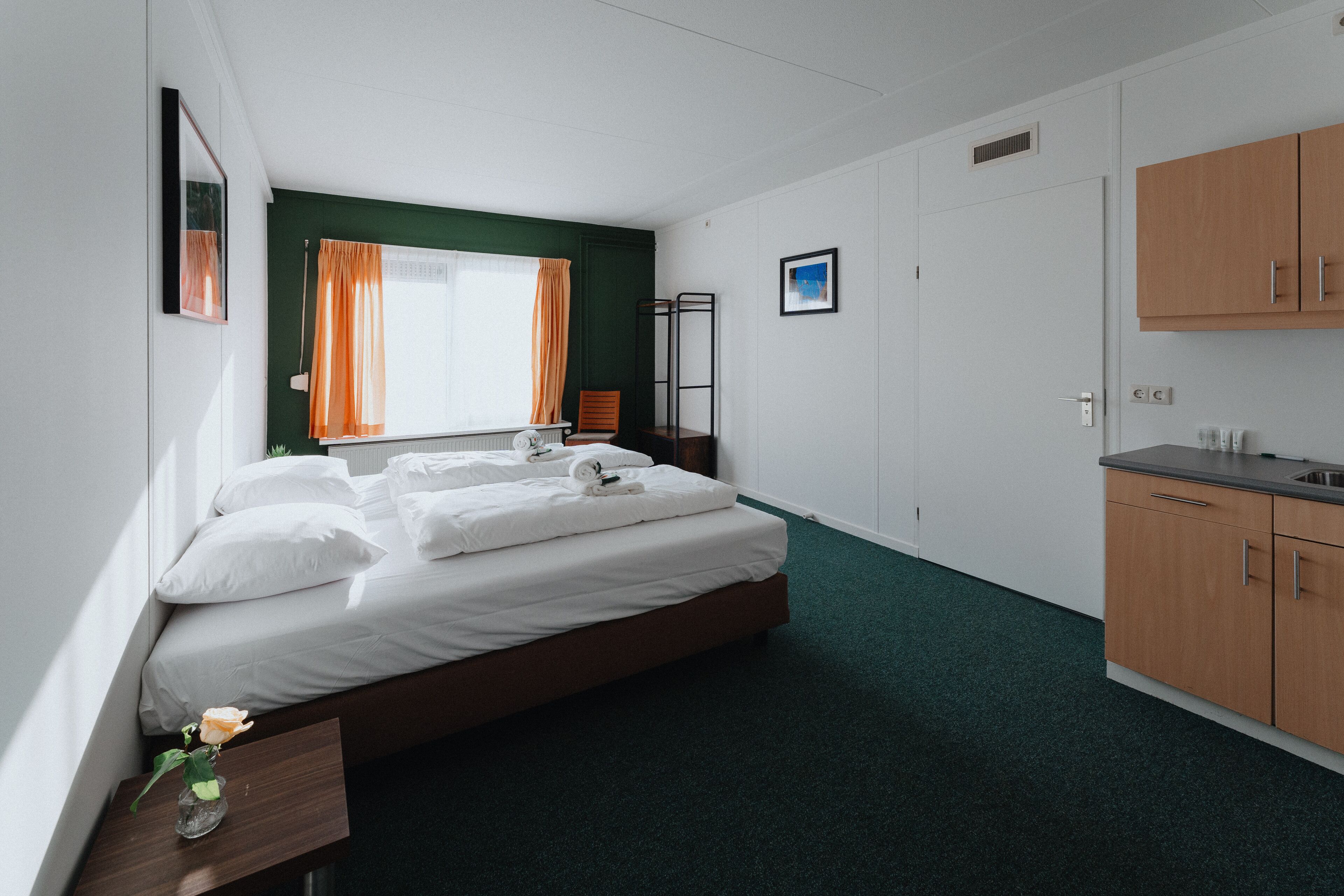 Habitación de hotel con cama, mesita de noche, ventana con cortinas, radiador, armario de madera y una zona de kitchenette.