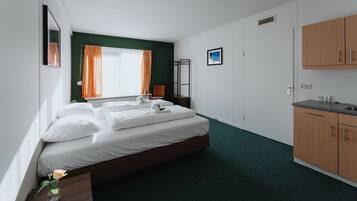 Habitación de hotel con cama, mesita de noche, ventana con cortinas, radiador, armario de madera y una zona de kitchenette.