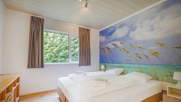 A bedroom with a large bed, a window with curtains, a wall mural of a beach, and a wooden nightstand with a lamp. - Landgoed Leudal (Haelen)
