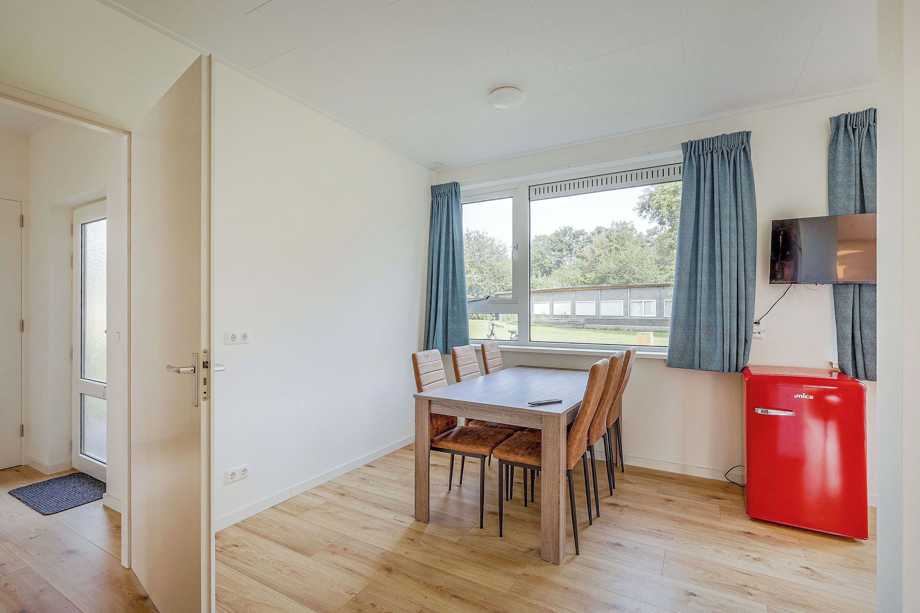 Une pièce comprenant une table à manger en bois, des chaises, un mini-réfrigérateur rouge, un téléviseur fixé au mur et une fenêtre avec des rideaux.